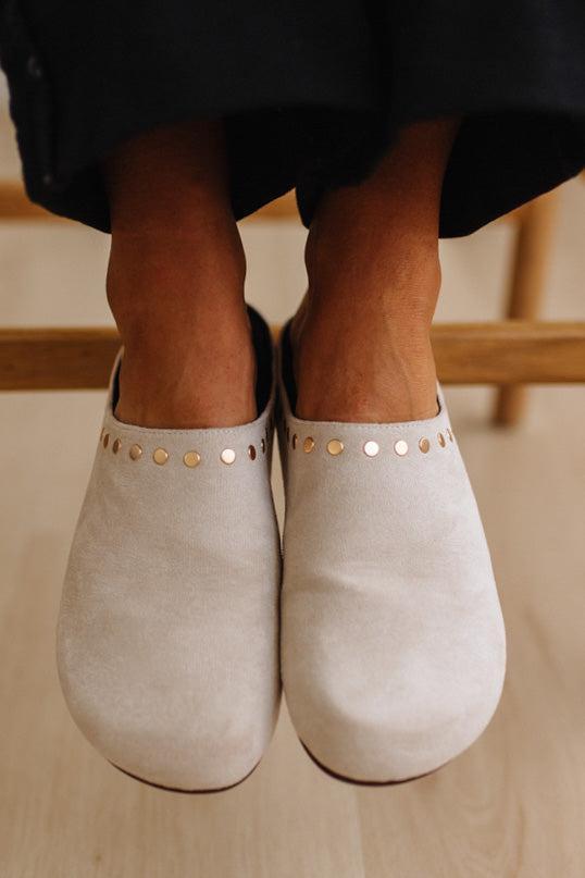 A close-up of a person wearing light beige slip-on clogs with metallic stud detailing around the collar, standing on a light wood floor.