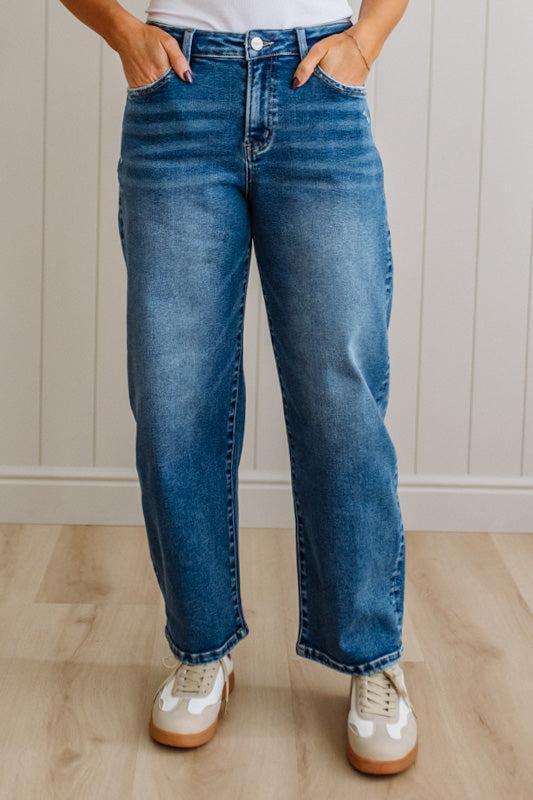 Person wearing blue jeans and white sneakers standing against a light wooden panel background