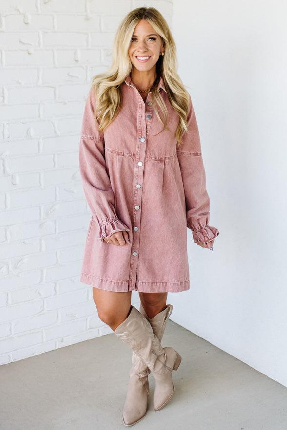 Woman wearing a pink denim dress with long sleeves against a white brick wall.