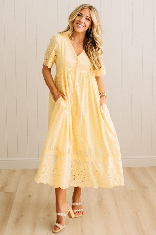 Woman wearing a yellow lace dress standing in a room with wooden flooring and white walls.