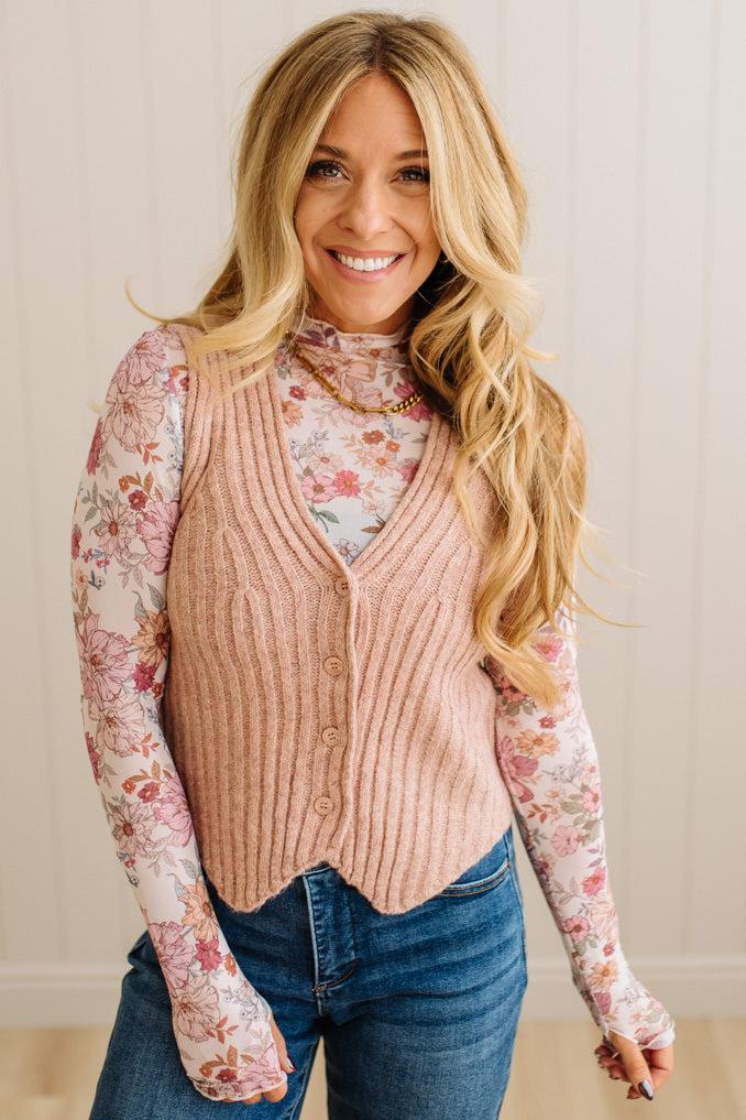 Blonde woman in dark jeans wearing a floral mesh top and ribbed sweater vest with button front.