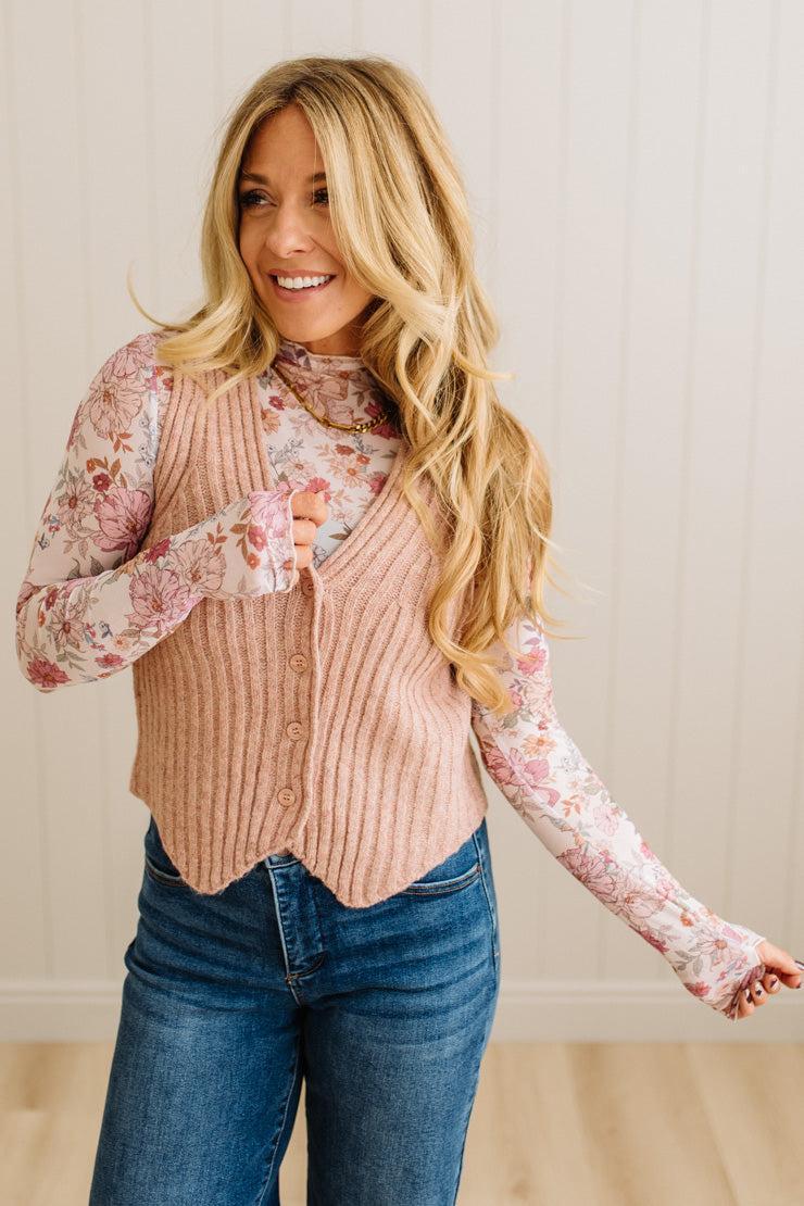 Woman wearing a pink ribbed vest over a floral blouse with blue jeans, standing against a neutral background.