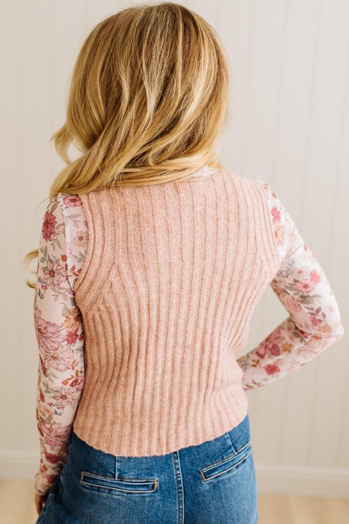 Person wearing a ribbed sweater vest over a floral shirt with blue jeans against a neutral background