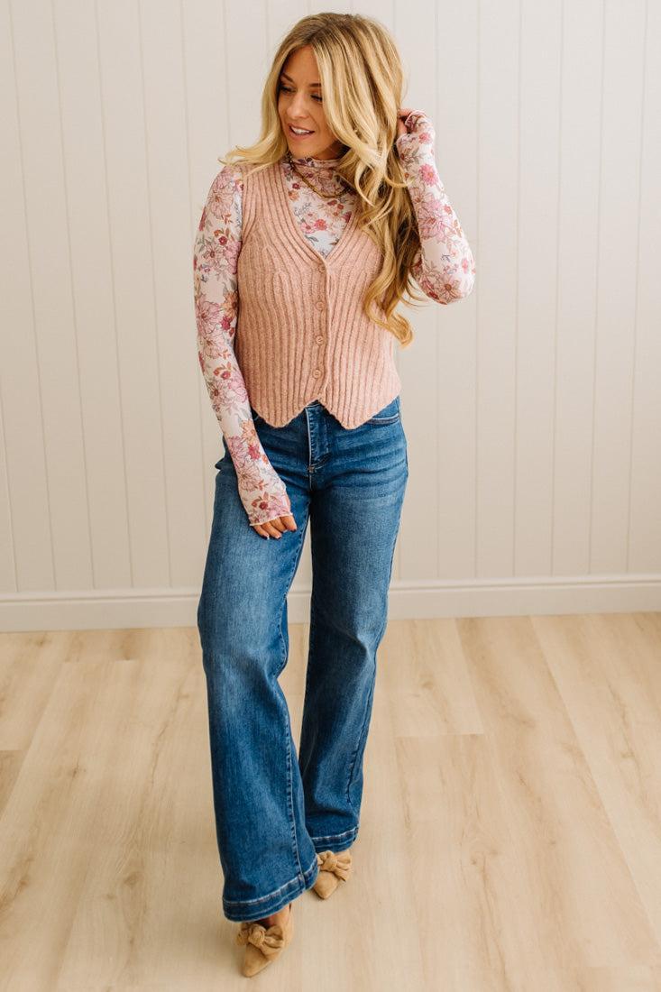 Woman wearing a pink ribbed sweater vest, floral blouse, and blue jeans on a wooden floor.