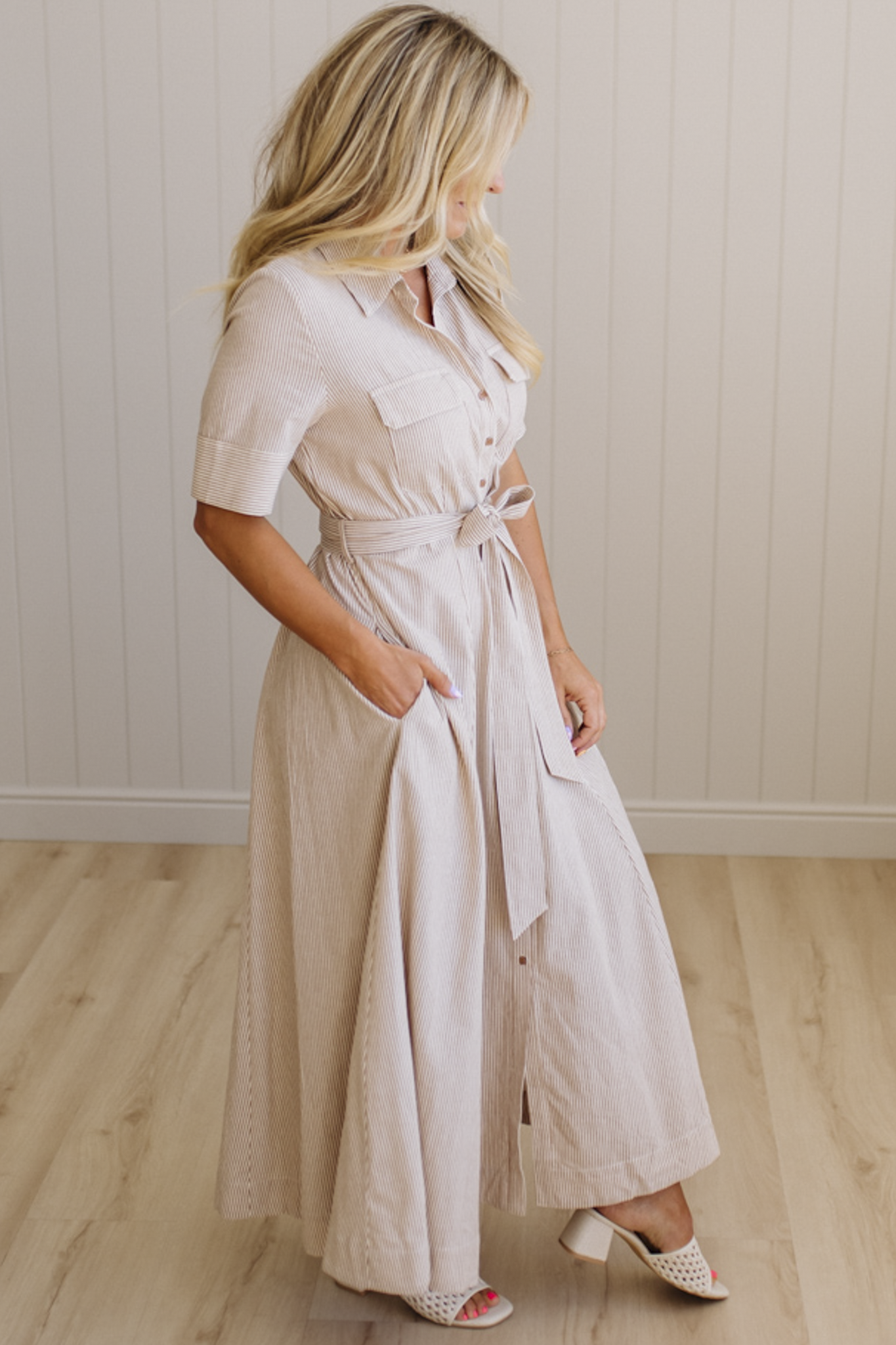 Woman wearing a long beige dress with a belt in a room with wooden flooring and a white wall.