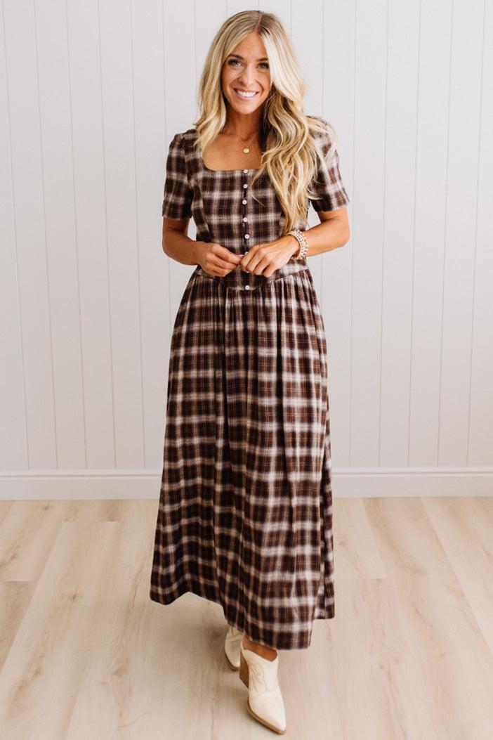 A woman modeling a brown and beige plaid midi dress with short sleeves, a square neckline, and a button-up front, standing in front of a white paneled wall and light wood floor.
