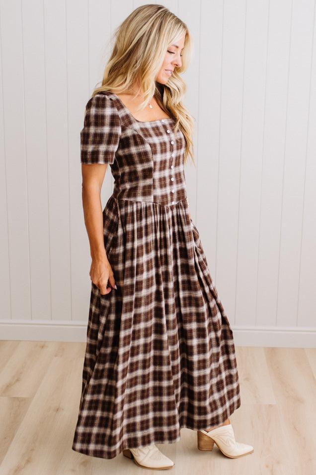 A woman modeling a brown and beige plaid maxi dress with short puff sleeves, a square neckline, and a gathered waist, standing in front of a white paneled wall and light wood floor.