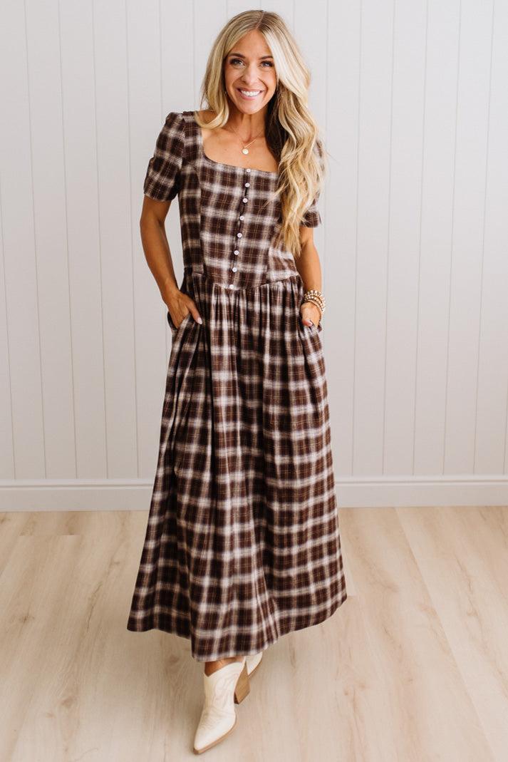 A woman modeling a brown and beige plaid maxi dress with short puff sleeves, a square neckline with button details, and side pockets, standing in front of a white paneled wall and light wood floor.