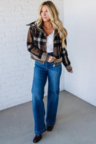 Woman wearing a plaid jacket, white shirt, and blue jeans against a white brick wall.