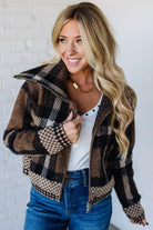 Woman wearing a plaid jacket over a white shirt and blue jeans against a white brick wall.