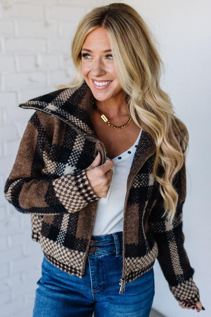 Woman wearing a plaid jacket over a white shirt and blue jeans against a white brick wall.