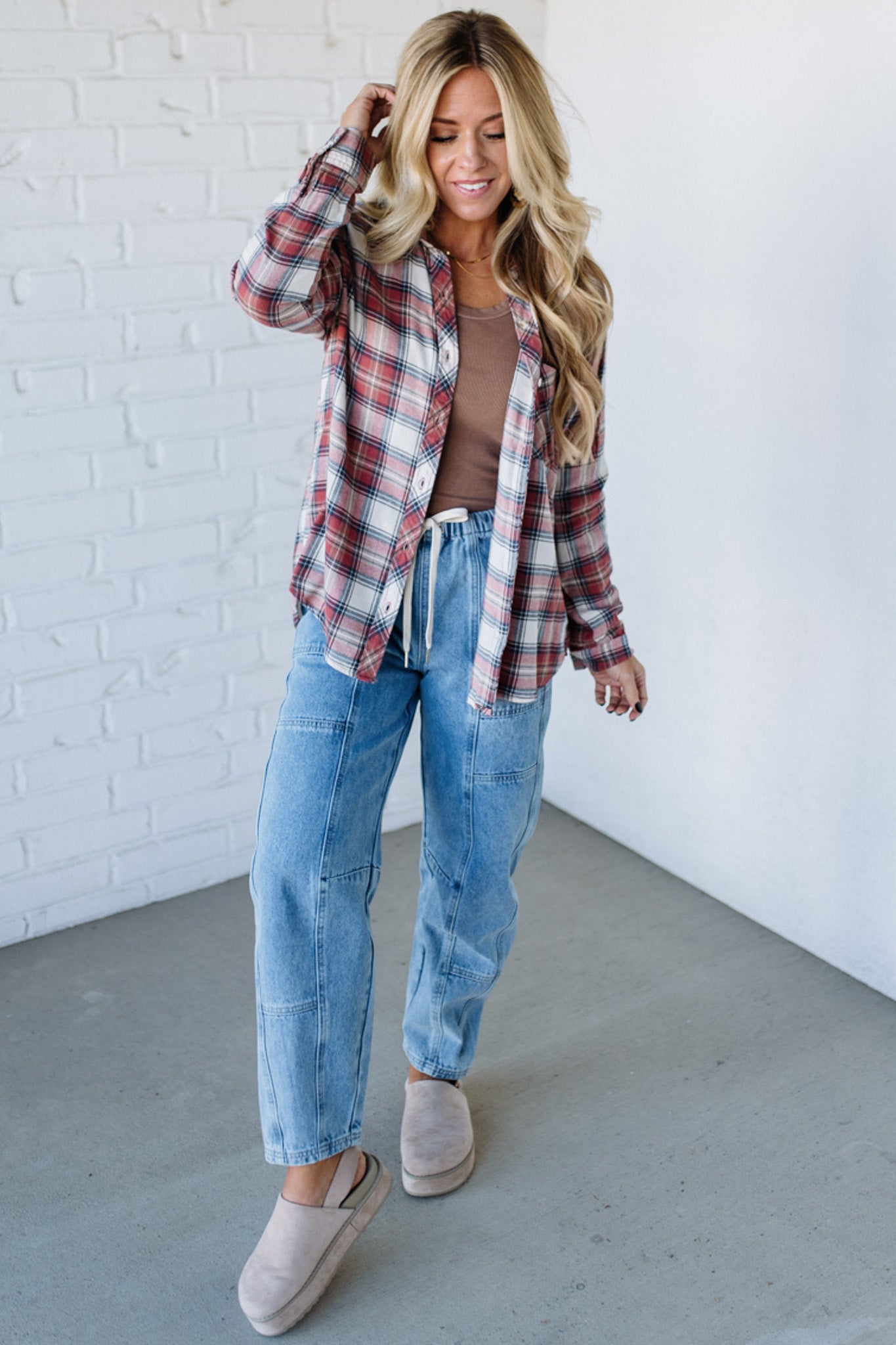 Woman wearing a plaid shirt, brown top, and blue jeans against a white brick wall.