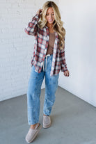 Woman wearing a plaid shirt, brown top, and blue jeans against a white brick wall.