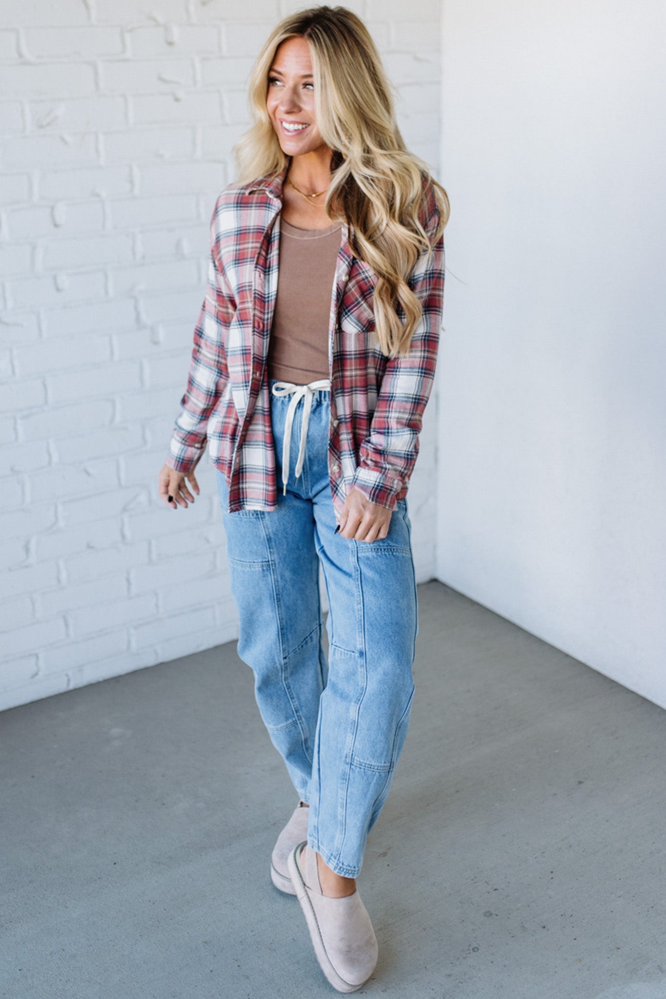 Woman wearing a plaid shirt, brown top, and blue jeans against a white brick wall.