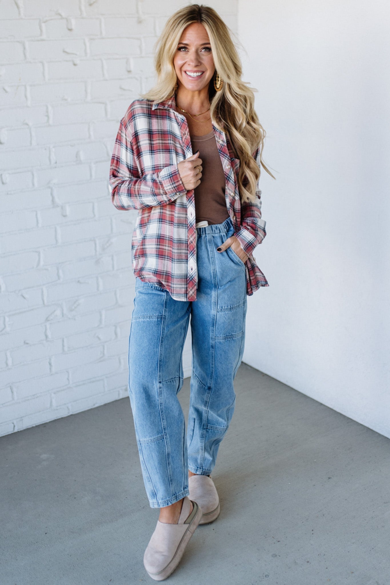Woman wearing a plaid shirt, jeans, and beige shoes against a white brick wall.