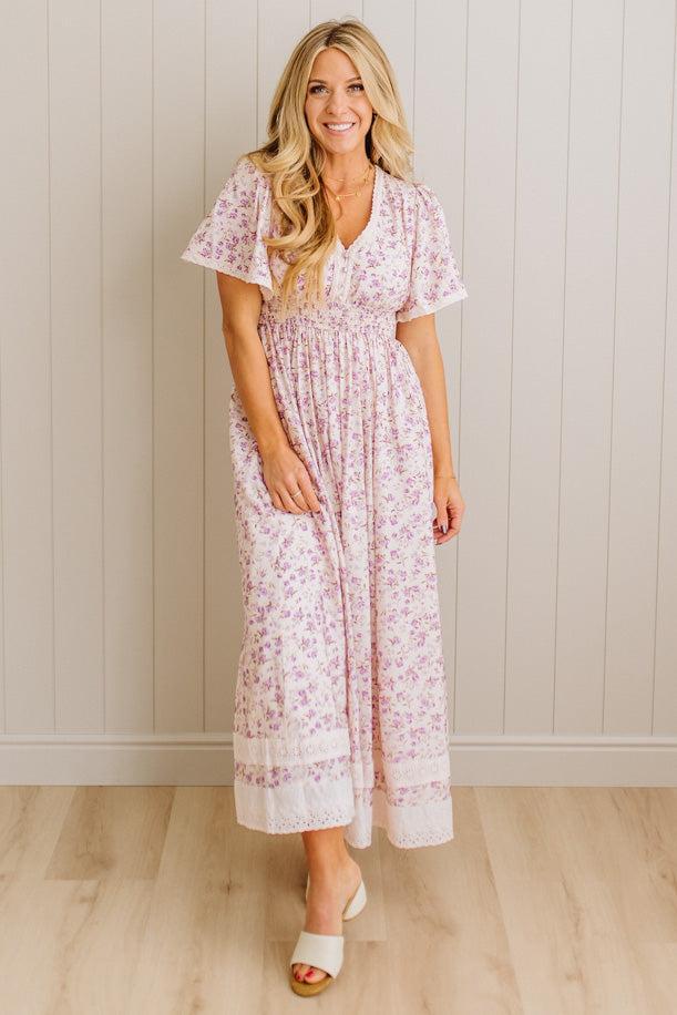 Woman wearing a floral dress standing against a light wooden paneled wall.