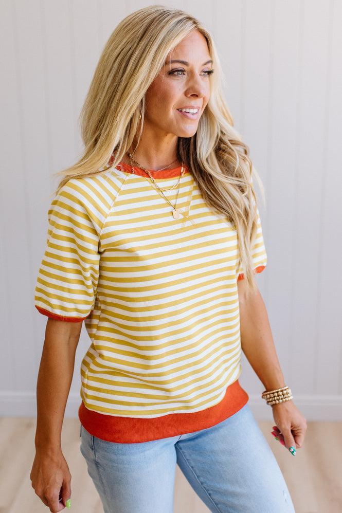 Blonde woman wearing light jeans and a mustard yellow with white stiripe short sleeve top, that also has a dark orange contrast trim.