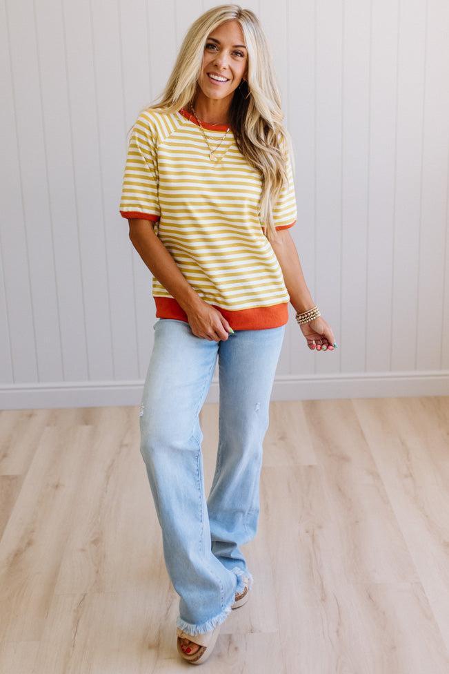Blonde woman wearing light jeans and a mustard yellow with white stiripe short sleeve top, that also has a dark orange contrast trim.