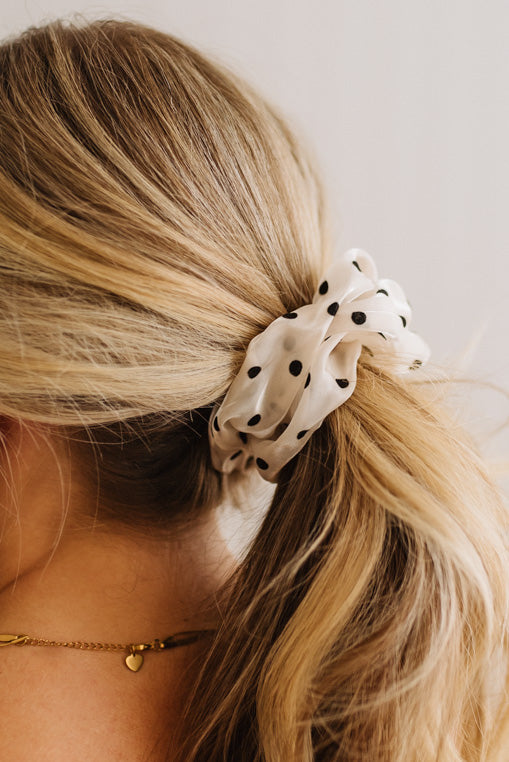 A close-up of a blonde woman's ponytail secured with a cream-colored fabric scrunchie featuring black polka dots, shown from the side against a light neutral background.