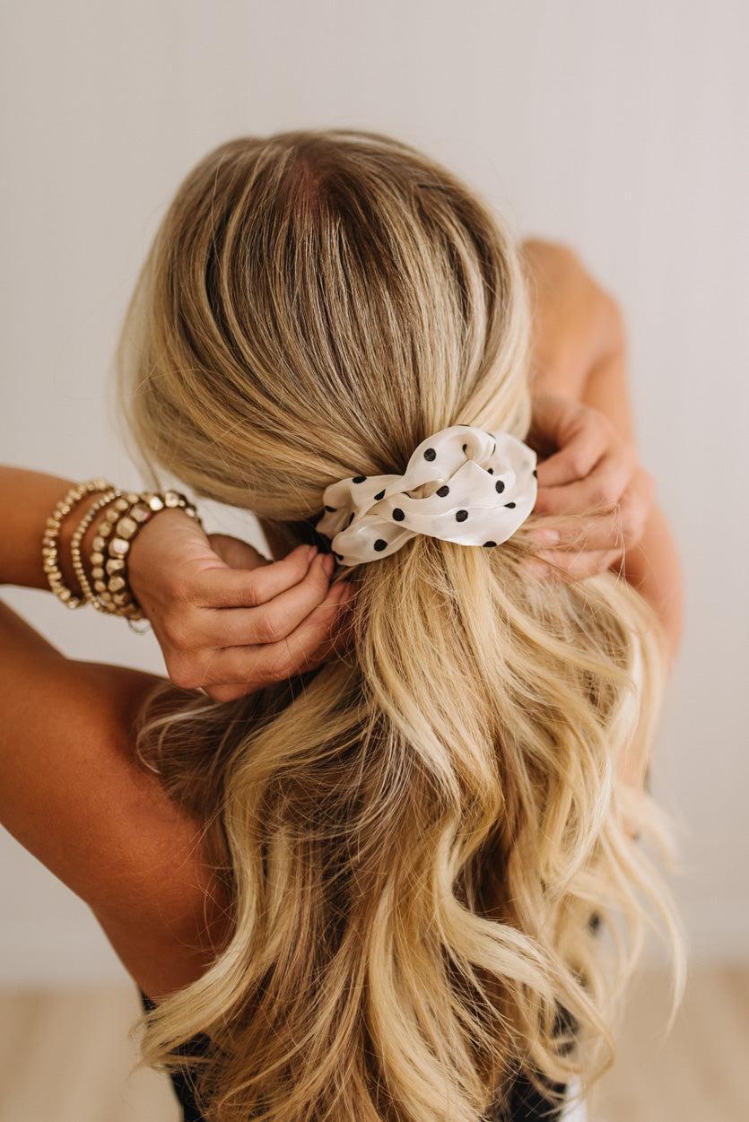 A blonde woman viewed from behind as she adjusts a white fabric scrunchie with black polka dots on her low ponytail. She is wearing stacked gold beaded bracelets against a plain light background.