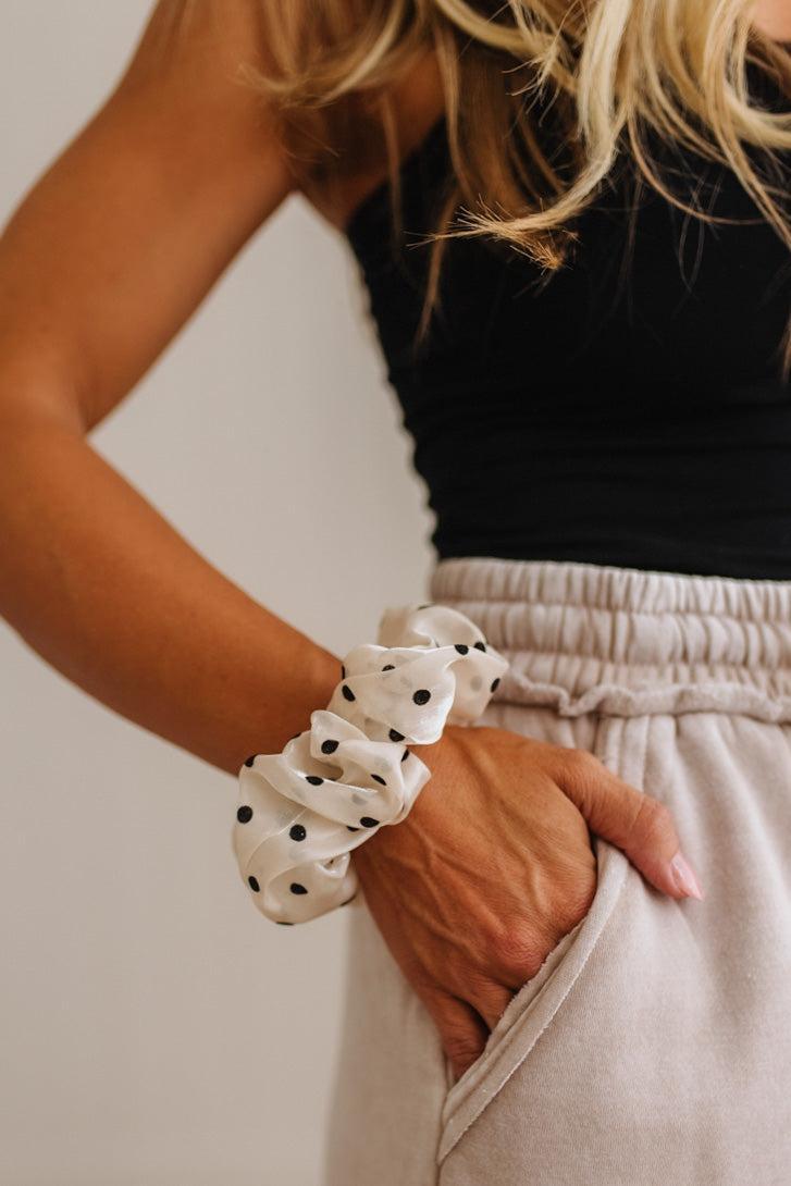 A close-up of a woman wearing a white fabric scrunchie with black polka dots, paired with a black top and beige sweatpants.