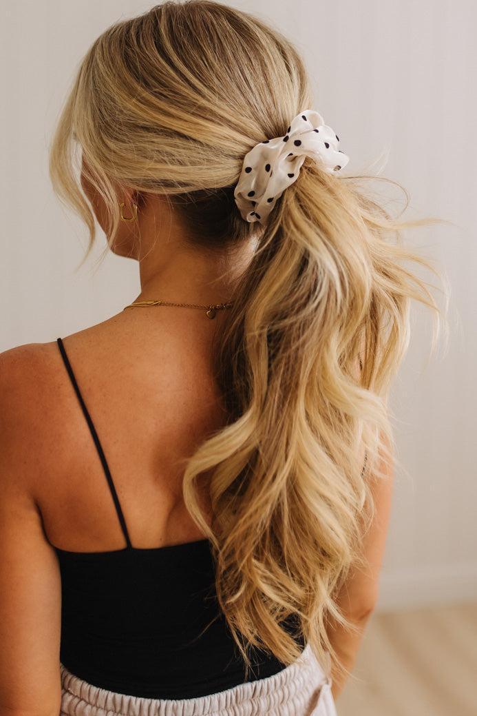 A woman with long blonde wavy hair seen from the back, wearing a white fabric scrunchie with black polka dots to hold her hair in a ponytail.