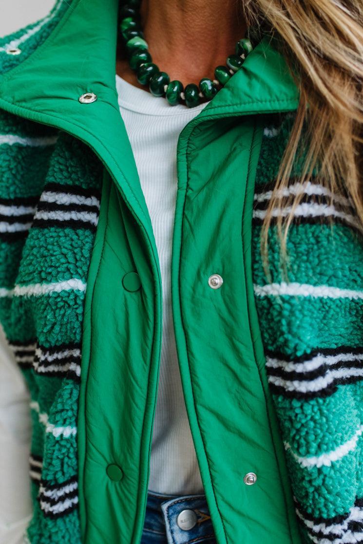 A close-up of a person wearing a bright green sherpa-lined puffer jacket with green, black, and white striped fleece panels, layered over a white top and blue jeans.