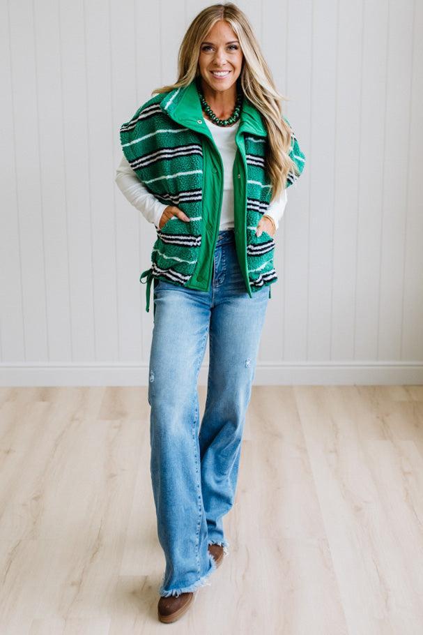 BLONDE WOMAN IN A STUDIO WITH LIGHT WOOD FLOORS AND LIGHT BACKDROP WEARING BLUE JEANS, A LONG SLEEVE WHITE TEE SHIRT AND A GREEN BLACK AND WHITE SHERPA COMFORT STRIPED VEST WITH SNAP BUTTONS AND SIDE POCKETS