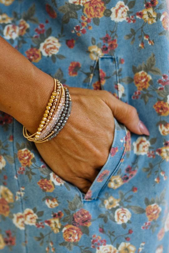 A close-up of a person's hand in a floral blue pocket, wearing a stack of beaded wrap bracelets in gold, silver, and rose gold tones.