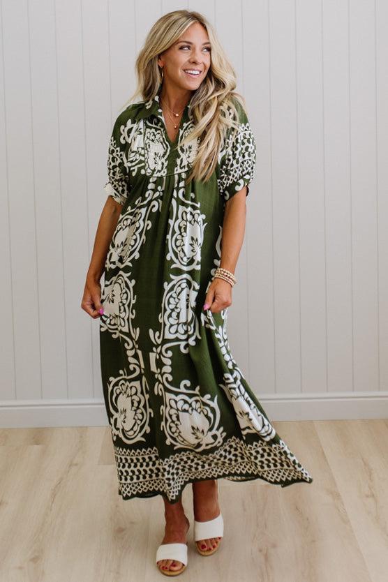 Woman wearing a green and white patterned dress against a white paneled wall.