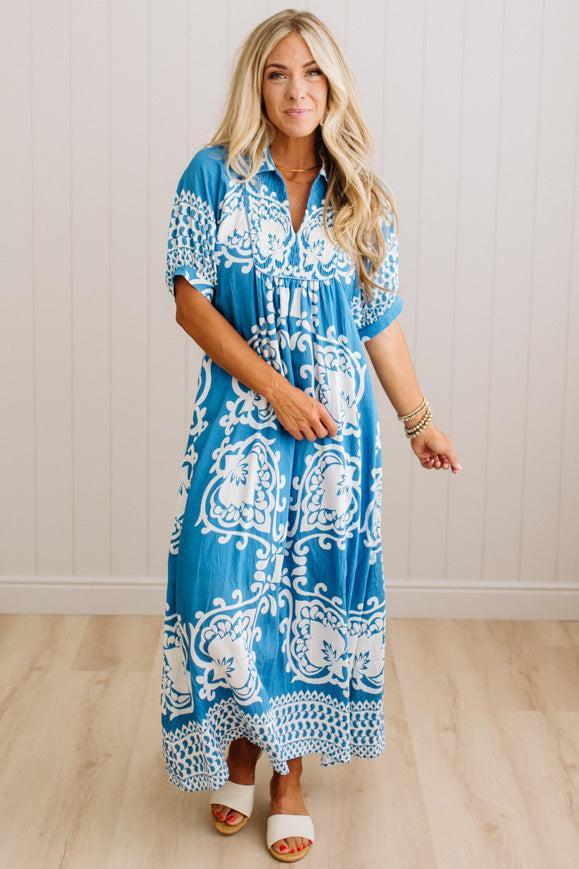 A woman modeling a blue and white patterned maxi dress with short sleeves and a V-neckline, standing against a white paneled wall and light wood floor.