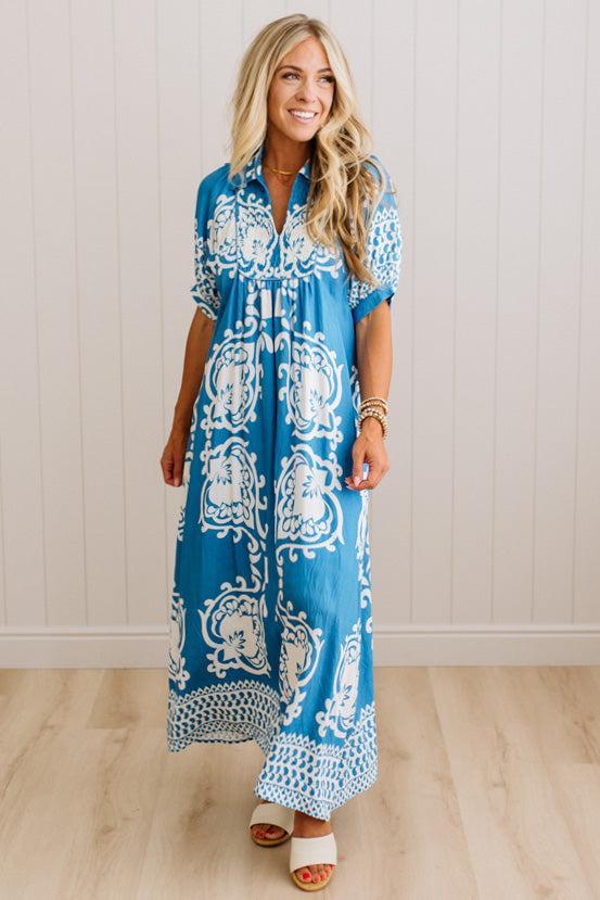 A woman modeling a blue and white patterned maxi dress with short sleeves, a tie neckline, and a loose, flowy silhouette, standing in a bright indoor setting with light wood floors and white paneled walls.