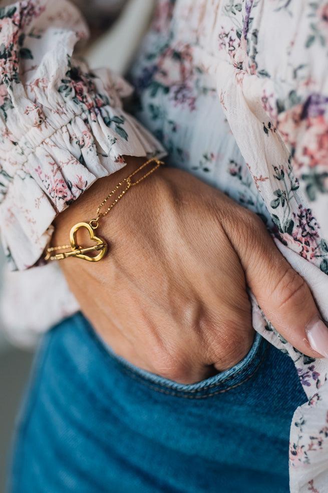 Hand wearing a gold bracelet with a heart charm, against a floral-patterned sleeve.