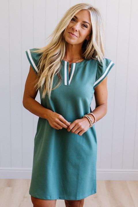 Woman wearing a teal dress with white trim against a white wall.