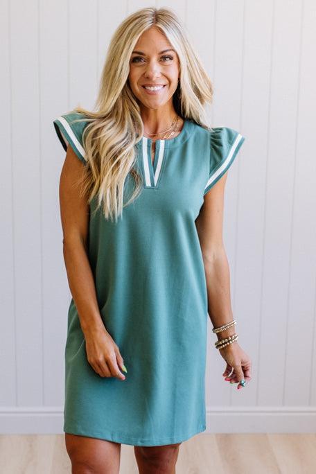 Woman wearing a teal dress with white trim against a white wall.