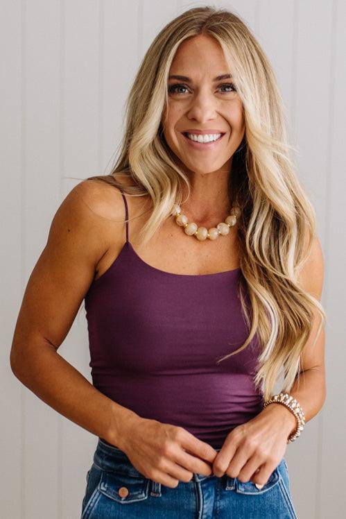 A smiling blonde woman wearing a fitted sleeveless camisole top in deep plum purple with blue jeans, standing against a light gray paneled wall.