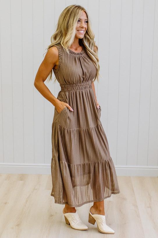 A woman modeling a sleeveless, tiered brown gingham maxi dress with side pockets, paired with cream-colored heeled mules, standing against a white paneled wall and light wood floor.
