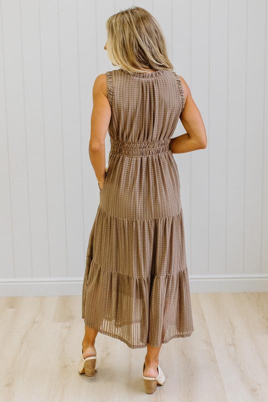 A woman modeling a sleeveless, tiered brown gingham maxi dress with side pockets, paired with white pointed-toe boots, standing against a white paneled wall and light wood floor.