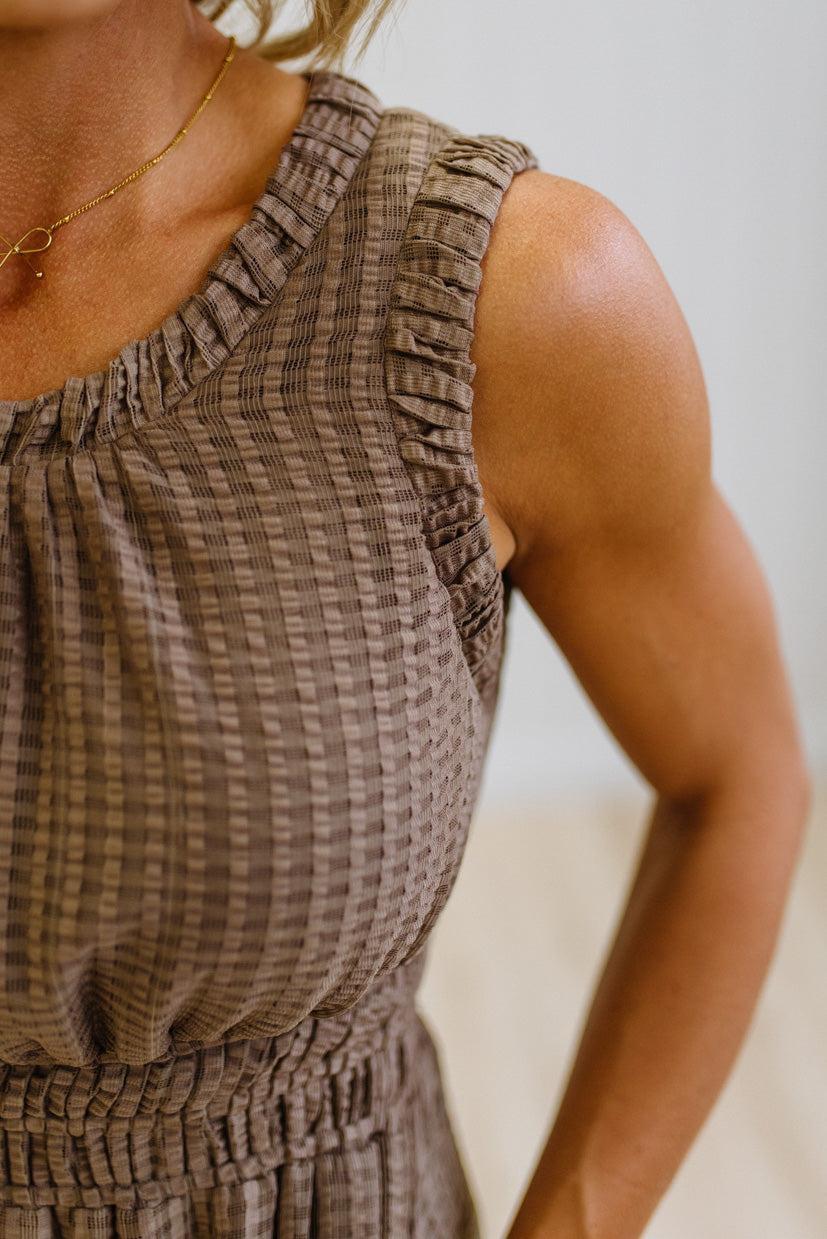 A woman modeling a sleeveless, tiered brown gingham maxi dress with side pockets, paired with white pointed-toe boots, standing against a white paneled wall and light wood floor.