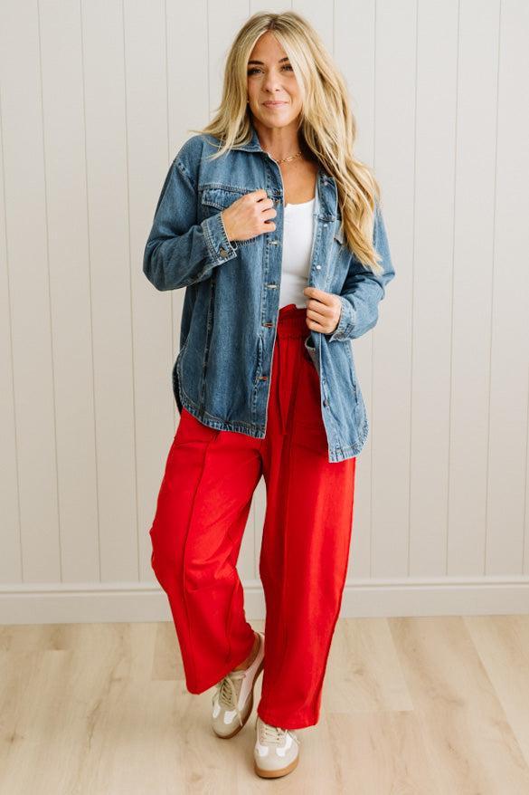 Woman wearing a denim jacket, white top, and red pants against a light wooden panel background