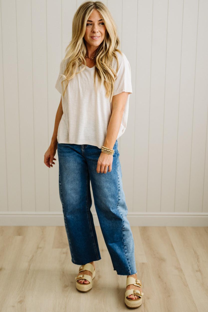 Woman wearing a cream t-shirt and blue jeans standing against a white paneled wall.