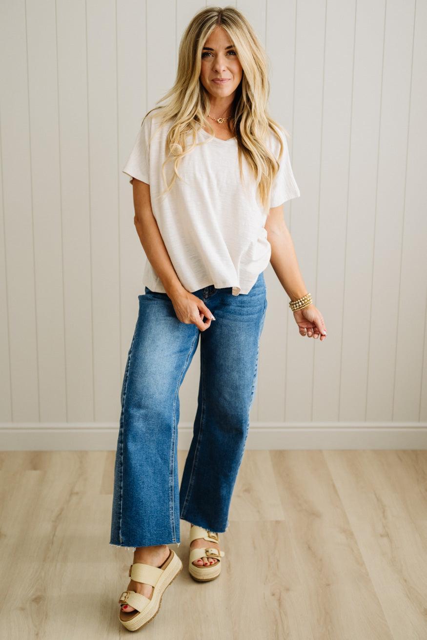 Woman wearing a sand beige t-shirt and blue jeans standing in a room with a light wooden floor and white walls.
