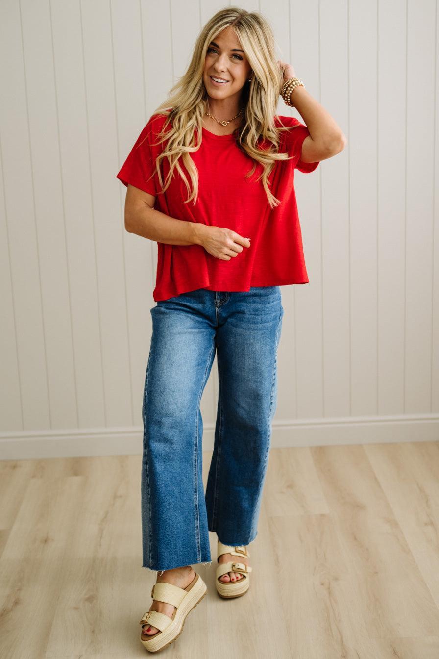Woman wearing a red top and blue jeans standing against a light wooden paneled wall.
