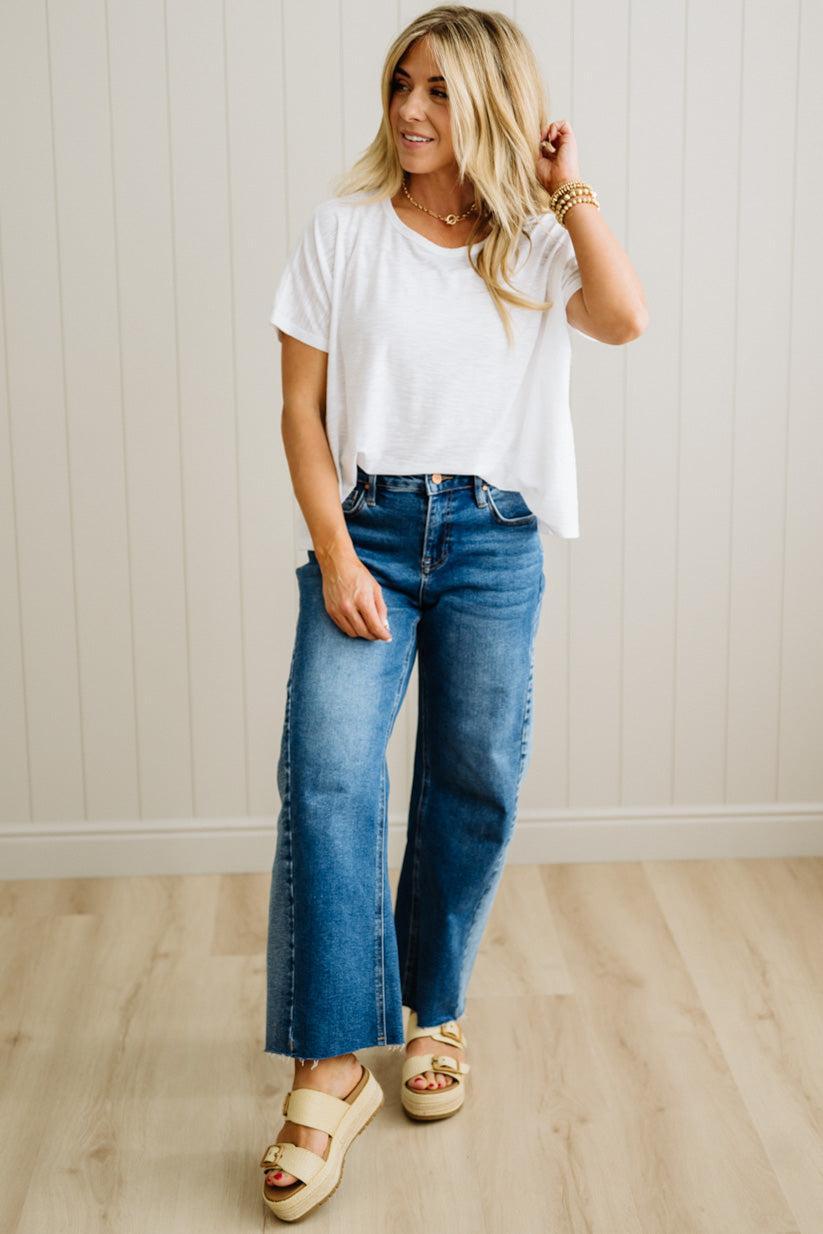 Woman wearing a white t-shirt and blue jeans standing against a light wooden paneled wall.