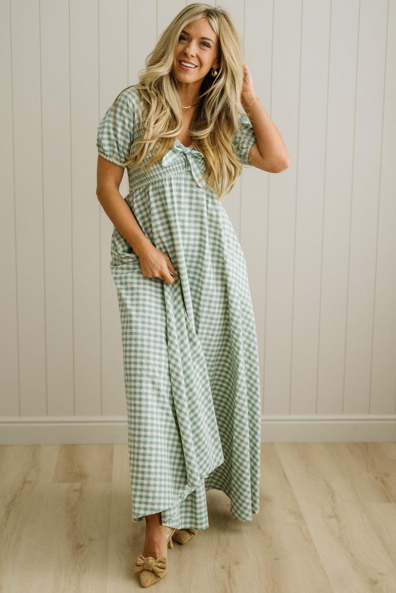 Woman wearing a green checkered dress standing against a light wooden paneled wall.