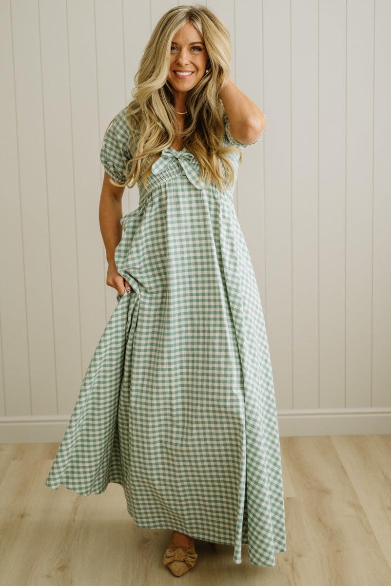Woman wearing a green checkered dress standing against a white paneled wall.