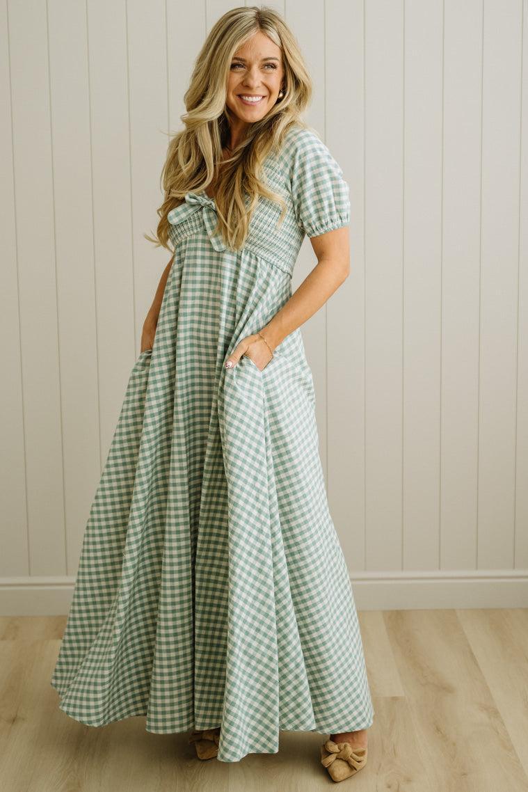 Woman wearing a green and white checkered dress standing against a light wooden paneled wall.