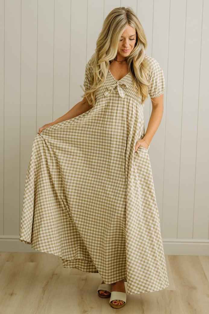 Woman wearing a checkered dress standing in a room with wooden flooring and a white paneled wall.
