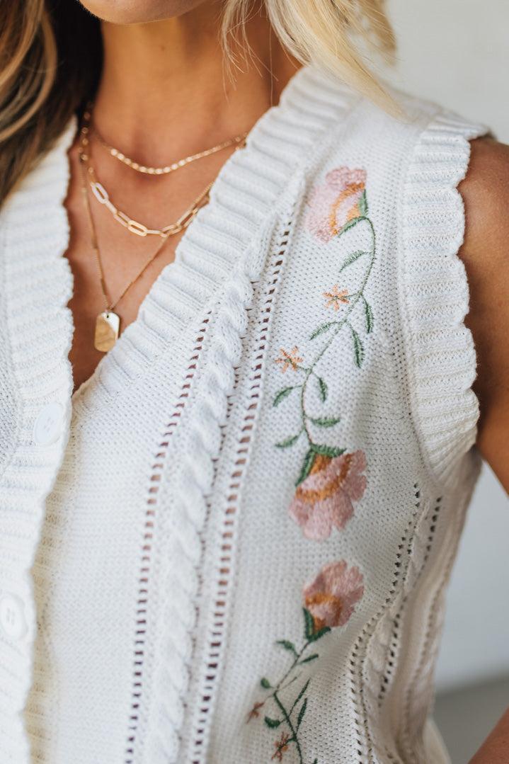 Blonde woman wearing a cream vest with scallop trim and floral embroidery.