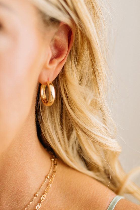 Close-up of a person wearing gold hoop earrings and a necklace with a neutral background